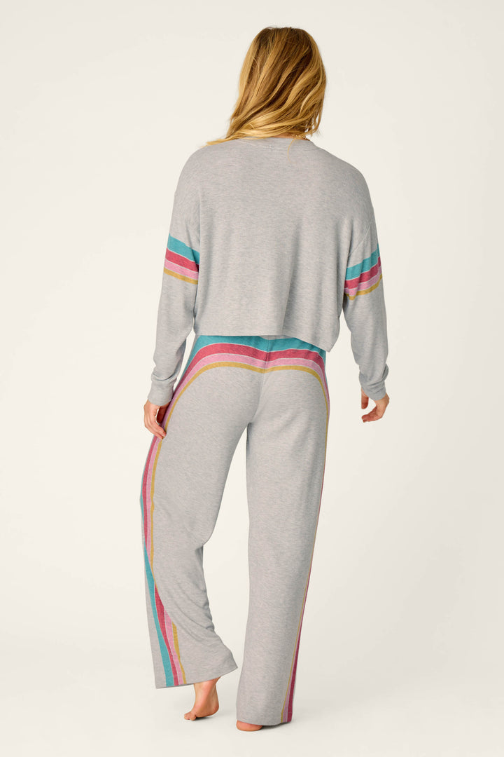 A person with long hair models the P.J. Salvage Takin’ It Easy Club Striped Lounge Set—a light gray set featuring colorful stripes along the arms and sides—standing barefoot and facing away from the camera against a plain background.