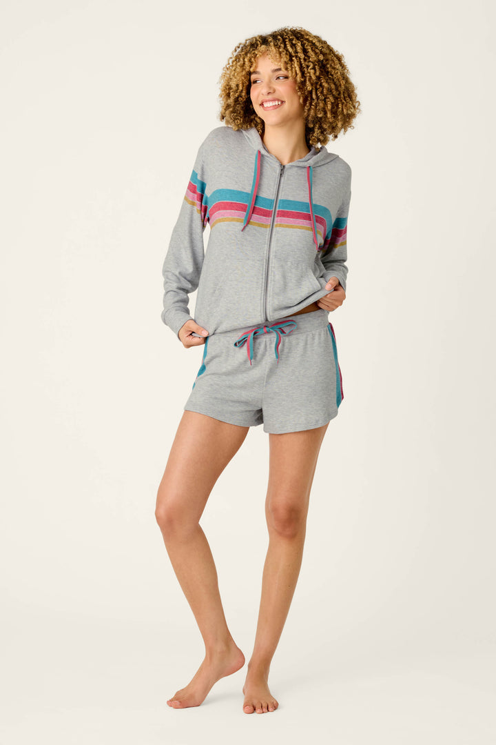 A person stands barefoot, smiling, wearing P.J. Salvage’s Takin’ It Easy Club Striped Shorts and a matching light gray zip-up hoodie with colorful stripes across the chest, arms, and sides, against a plain off-white background.