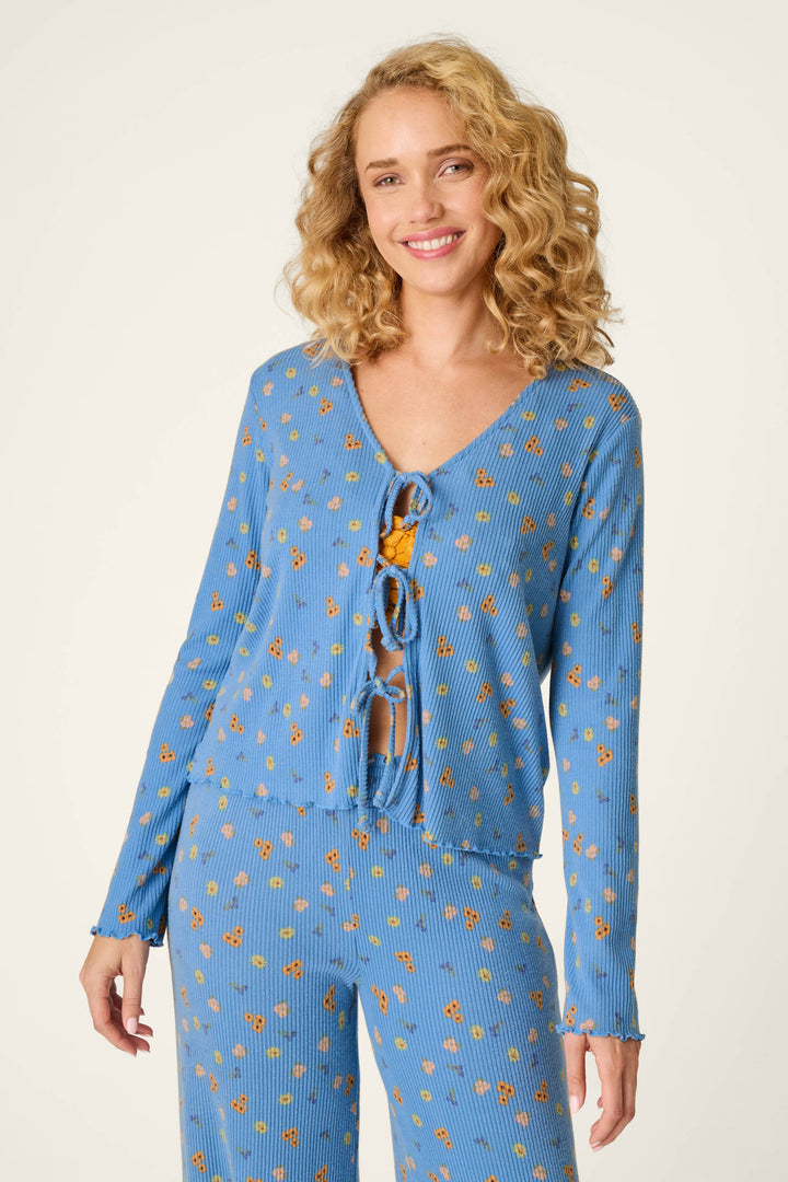 A smiling woman with curly blonde hair models the P.J. Salvage Tiny Blooms Paris Blue Floral Print Tie-Front Top, featuring long sleeves, three front ties, and a pattern of small orange flowers. She pairs it with matching pants and reveals a hint of an orange undergarment against a plain light background.