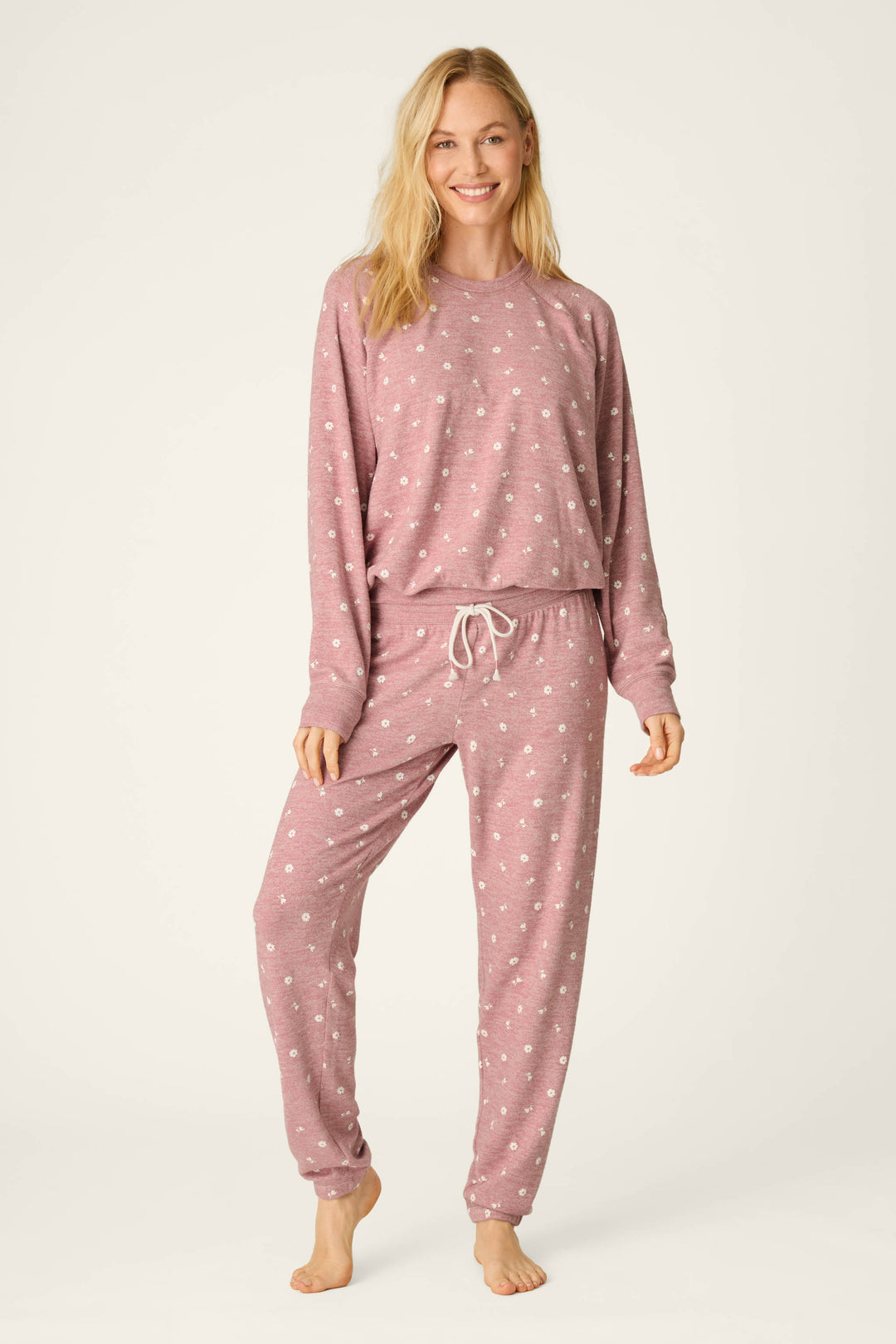 A woman smiles while wearing the Tiny Blooms Polka Dot Long Sleeve Top by P.J. Salvage, paired with matching drawstring pants in a pink and white polka dot pajama set. She stands barefoot against a plain, light background.