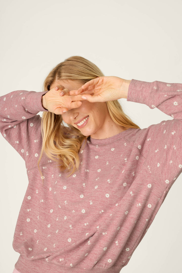 A person with long blonde hair smiles while covering part of their face with their hands. They are wearing the P.J. Salvage Tiny Blooms Polka Dot Long Sleeve Top, which features a pink color with a white floral pattern, and they stand in front of a plain, light background.
