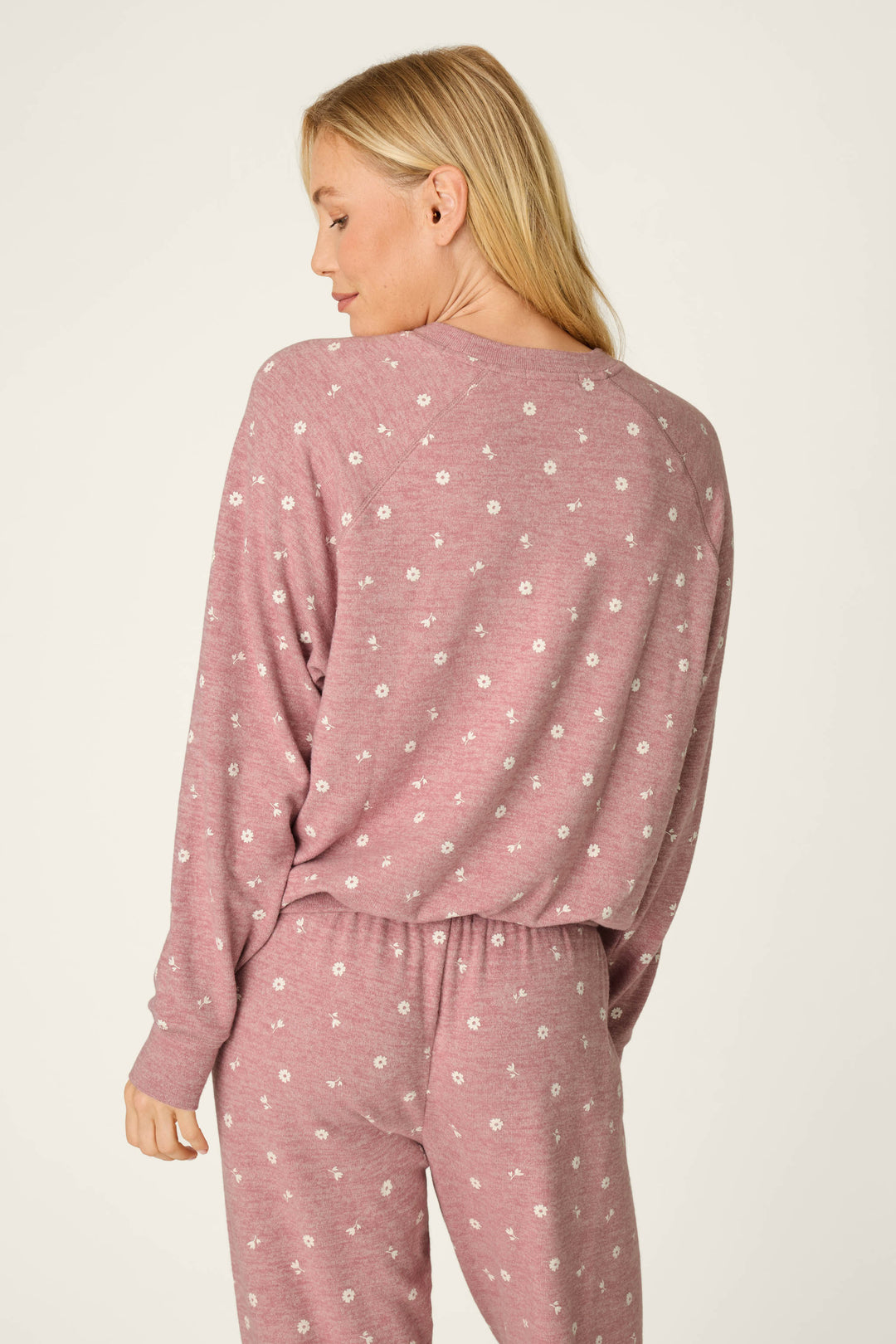 A woman with blonde hair models the P.J. Salvage Tiny Blooms Polka Dot Long Sleeve Top—a pink pajama top featuring small white floral patterns. She is turned slightly away, glancing over her shoulder with a relaxed expression against a simple, light-colored background.
