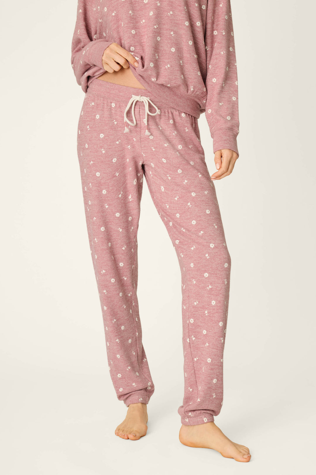 A person is wearing the P.J. Salvage Tiny Blooms Polka Dot Pajama Jogger Pants, featuring a light pink color with a white floral pattern and a drawstring waist. The image captures the body from the shoulders down, showing the individual standing barefoot against a light background.