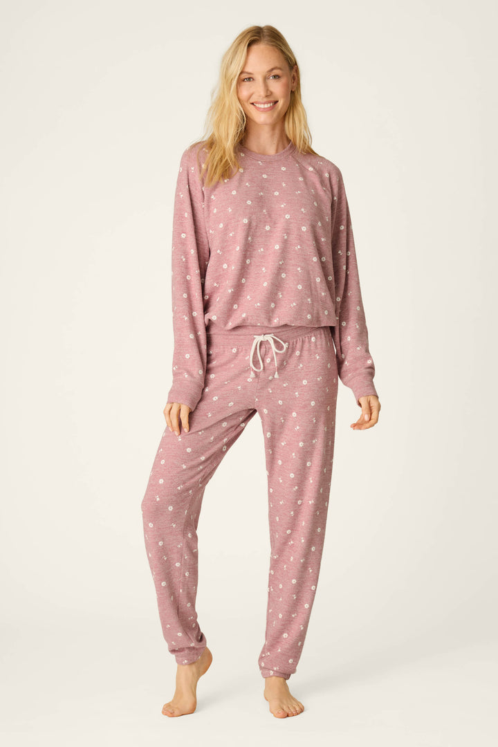A person stands barefoot against a plain light background, smiling with one hand by their side and the other touching their leg, wearing the P.J. Salvage Tiny Blooms Polka Dot Pajama Jogger Pants in a matching mauve set with small white polka dots.