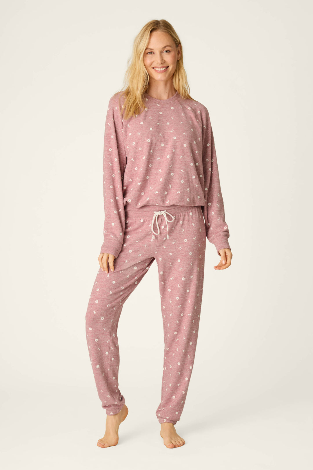 A woman stands barefoot against a light background, smiling with one hand on her hip, wearing the Tiny Blooms Polka Dot Long Sleeve PJ Set by P.J. Salvage—a matching pink pajama set with white polka dots.