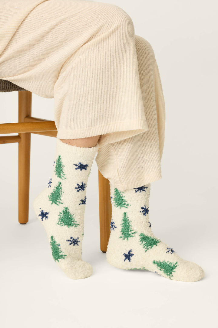 A person seated on a wooden chair wearing cream ribbed socks with a green tree and navy star pattern, paired with cream knit pants against a light neutral background.