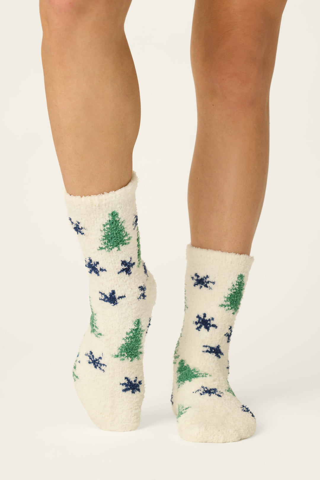 Close-up of a person's lower legs wearing P.J. Salvage's Under the Stars Tree & Star Print Socks—cozy white socks decorated with green trees and blue star motifs—standing on a light background.