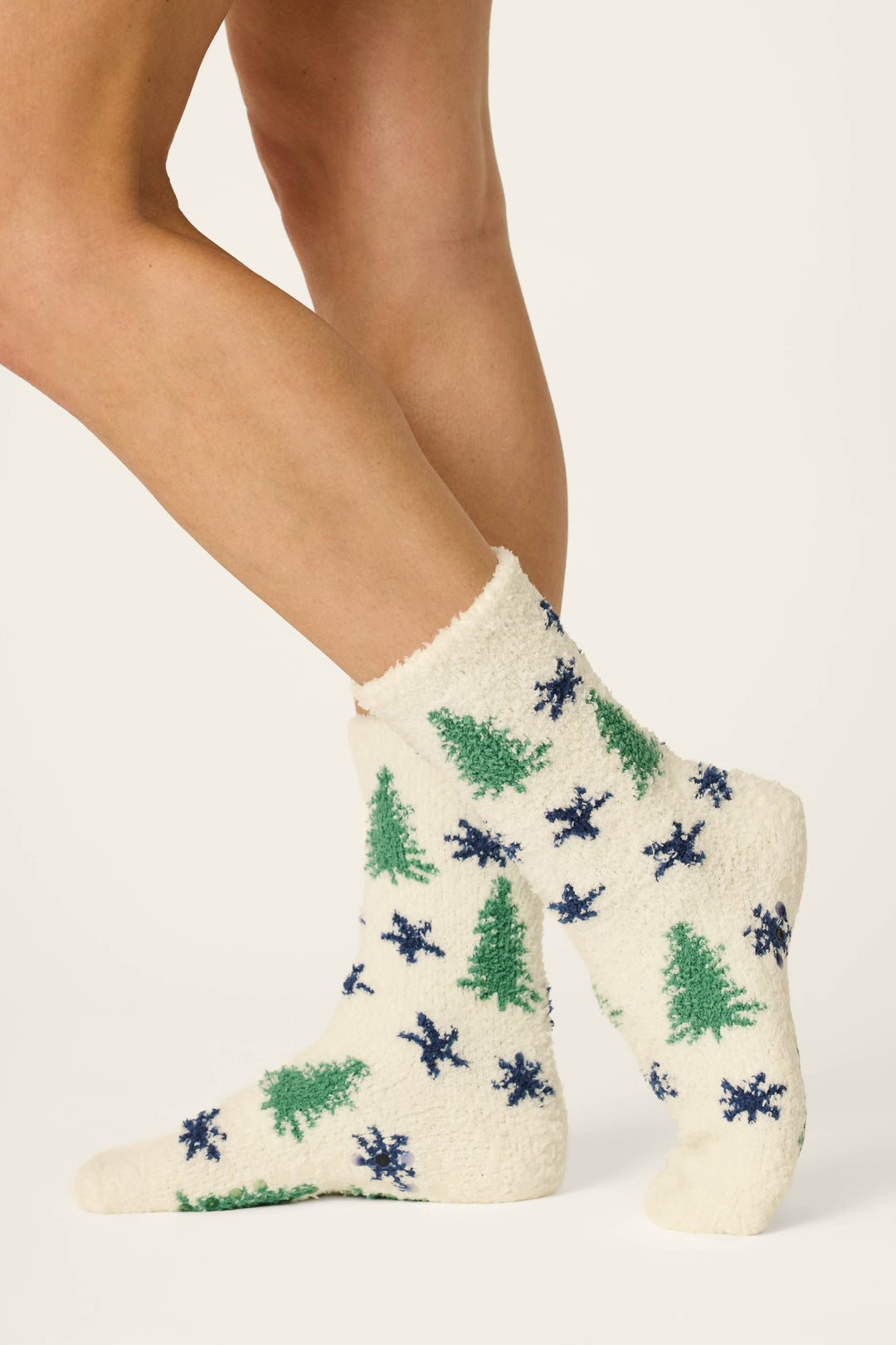 A person wears P.J. Salvage "Under the Stars Tree & Star Print Socks," cozy white socks featuring green Christmas trees and blue snowflakes, while standing on a plain white background.