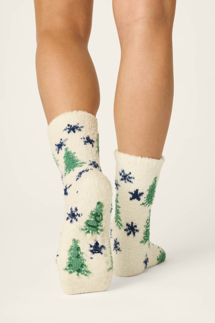 A person wearing P.J. Salvage's Under the Stars Tree & Star Print Socks, featuring fuzzy white fabric decorated with green Christmas trees and blue snowflakes, stands with their back to the camera against a plain background.