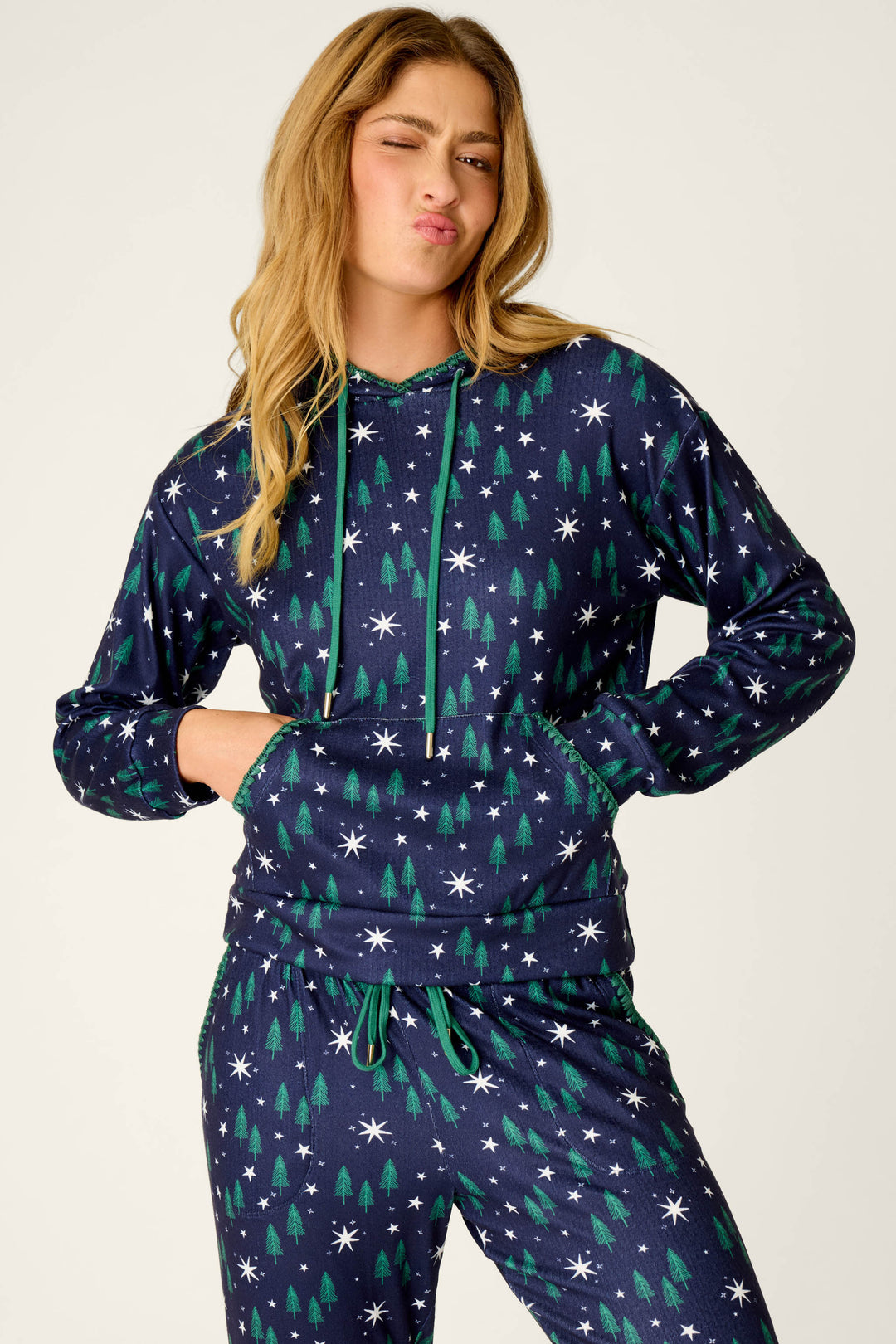 A woman wearing the P.J. Salvage “Under the Stars Magical Forest Print Hoodie,” featuring a navy base adorned with green tree and white star patterns, stands with her hands in her pockets while making a playful duck face.