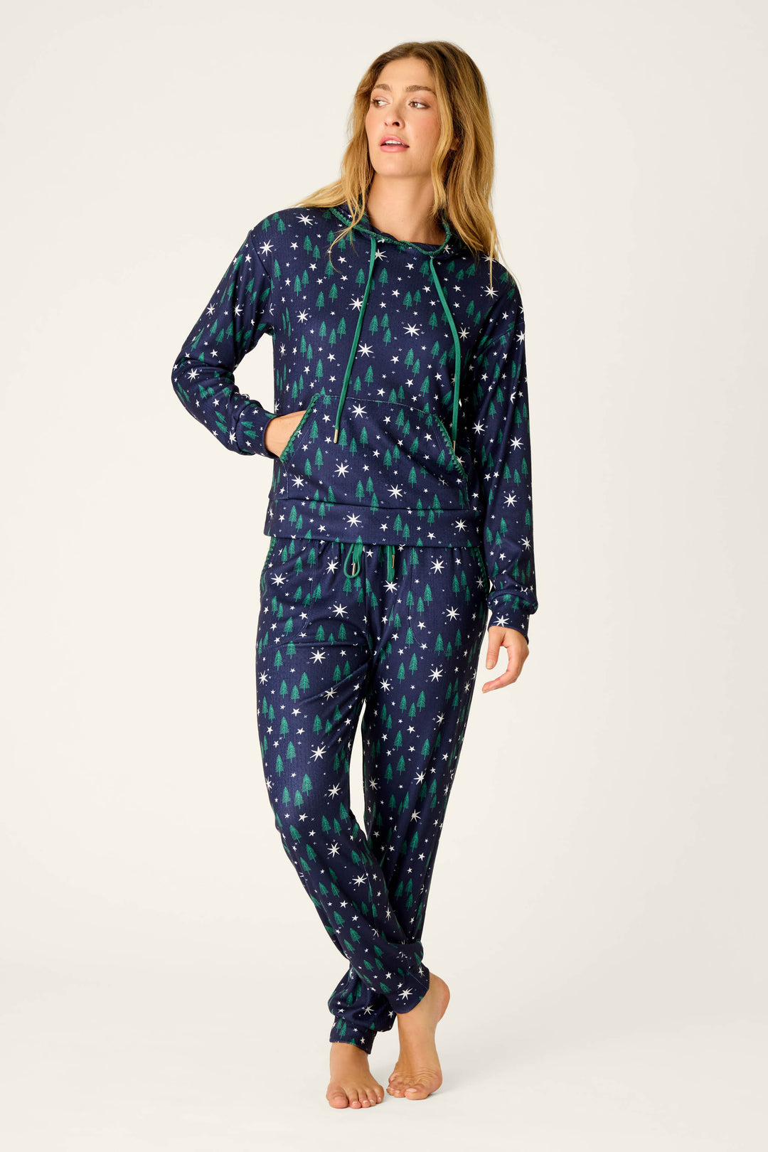 A woman wears the P.J. Salvage “Under the Stars Magical Forest Print Hoodie,” featuring a navy blue base with green and white tree and star patterns, standing barefoot against a plain light background.