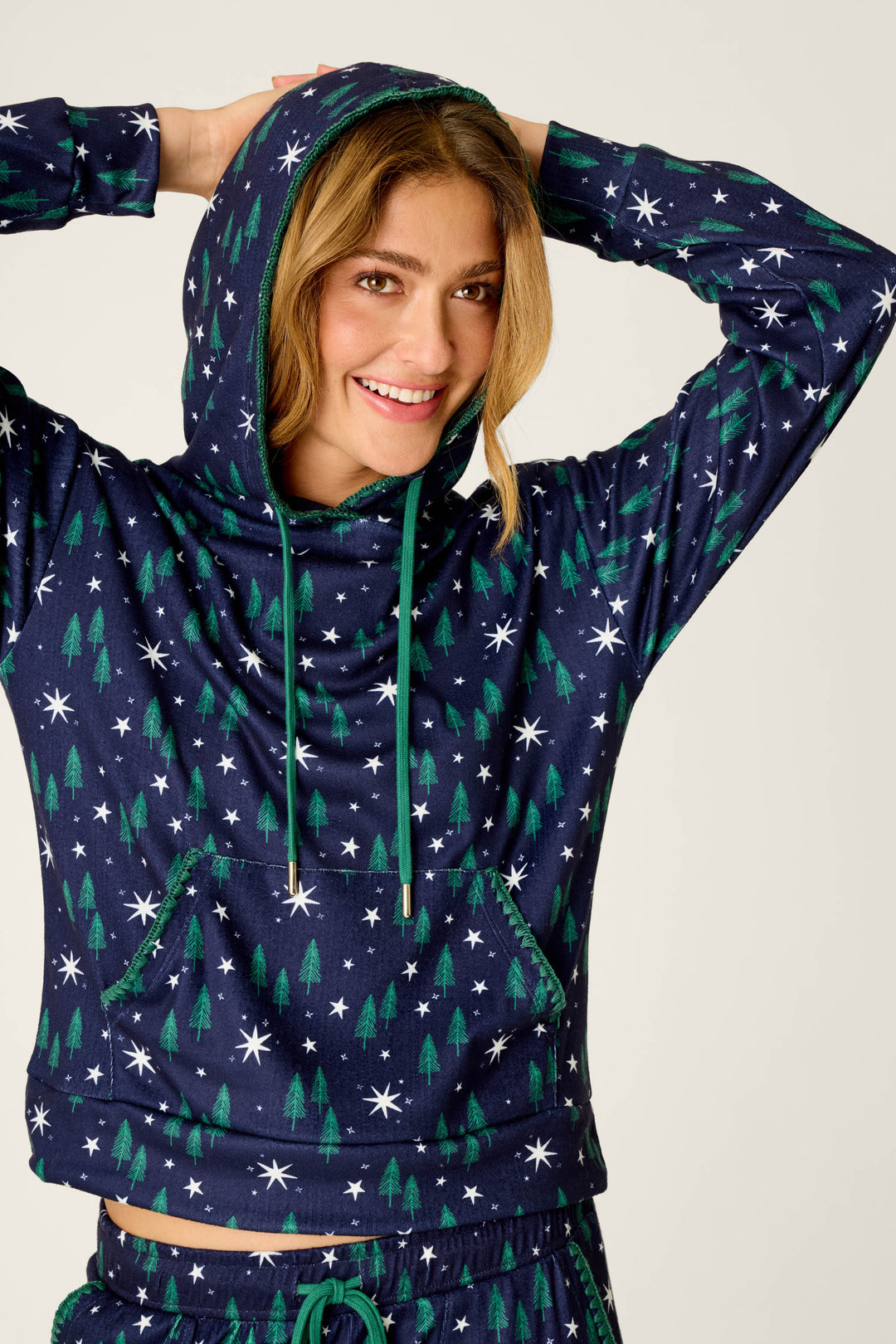A smiling person stands against a plain background in the P.J. Salvage Under the Stars Magical Forest Print Hoodie, featuring green trees and white stars on navy blue, with hands behind their head.