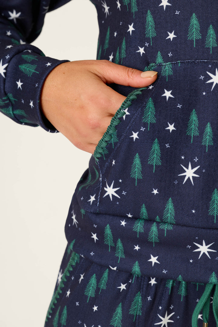 A person’s hand rests in the pocket of a P.J. Salvage “Under the Stars Magical Forest Print Hoodie,” which features a navy blue design with green trees and white star patterns.