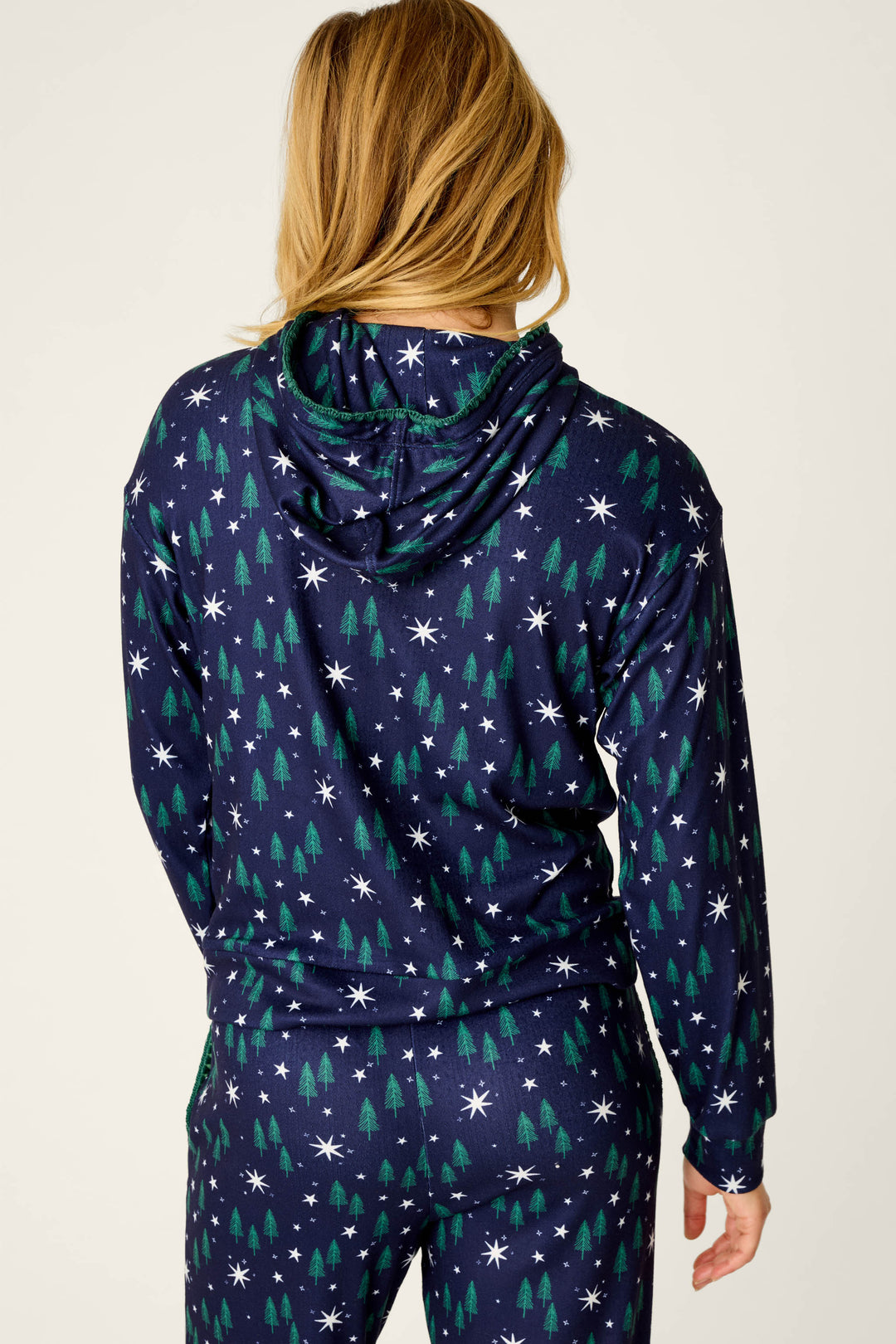 A person with long blonde hair stands with their back to the camera, wearing the P.J. Salvage Under the Stars Magical Forest Print Hoodie and matching pants set decorated with white stars and green trees.