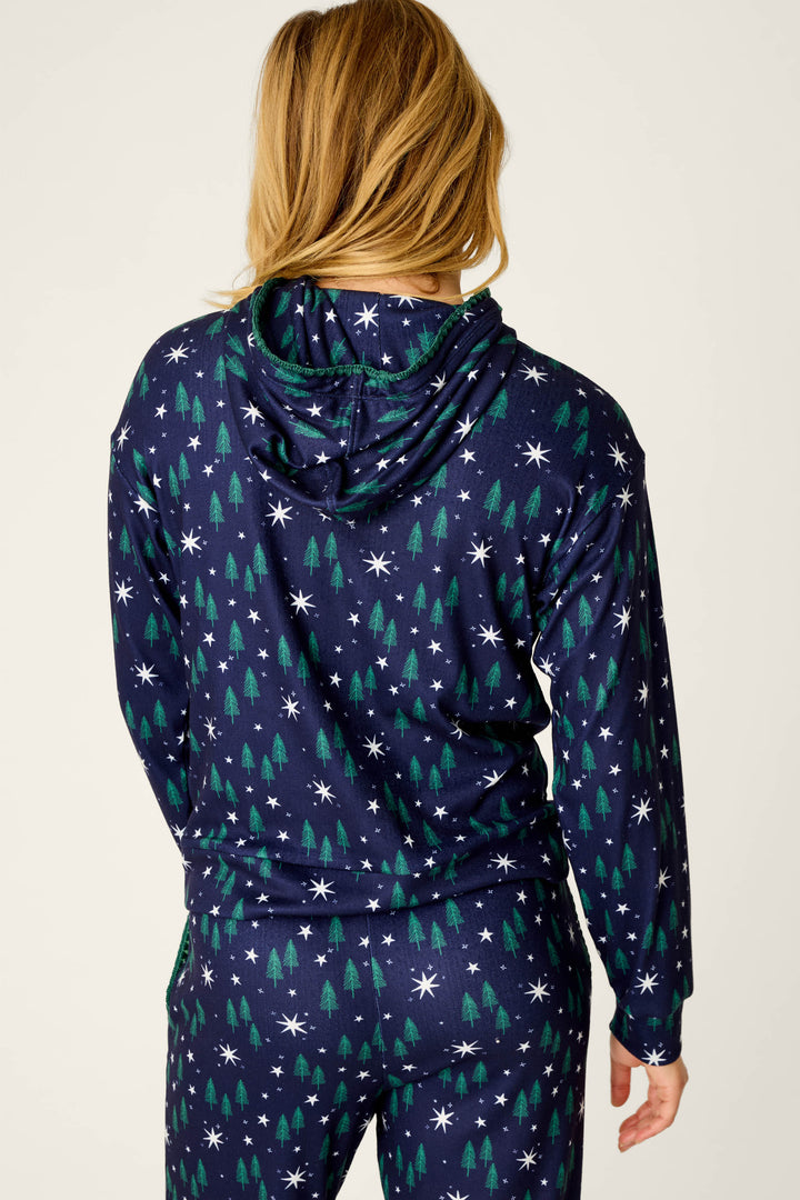 A person with long blonde hair stands with their back to the camera, wearing the P.J. Salvage Under the Stars Magical Forest Print Hoodie and matching pants set decorated with white stars and green trees.