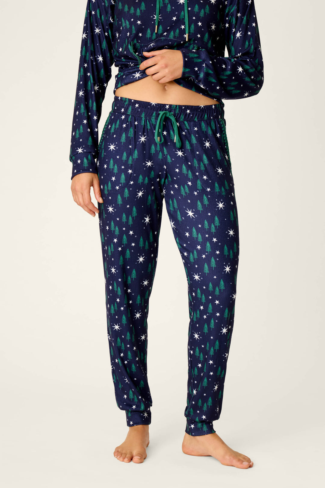 A person models the Under the Stars Magical Forest Print Joggers by P.J. Salvage, featuring a navy blue base with green tree and white snowflake patterns. The photo shows only their body from mid-chest to feet, standing barefoot against a plain background.