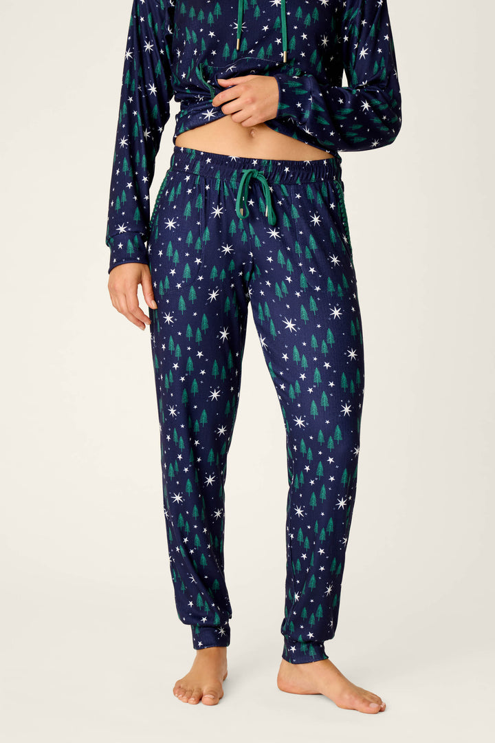 A person models the Under the Stars Magical Forest Print Joggers by P.J. Salvage, featuring a navy blue base with green tree and white snowflake patterns. The photo shows only their body from mid-chest to feet, standing barefoot against a plain background.