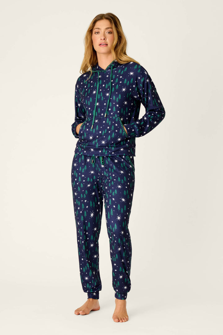 A woman stands barefoot against a plain light background, hands in her pockets, wearing the P.J. Salvage Under the Stars Magical Forest Print Joggers featuring a matching navy blue pajama set with a hood, decorated with green Christmas trees and white snowflakes.