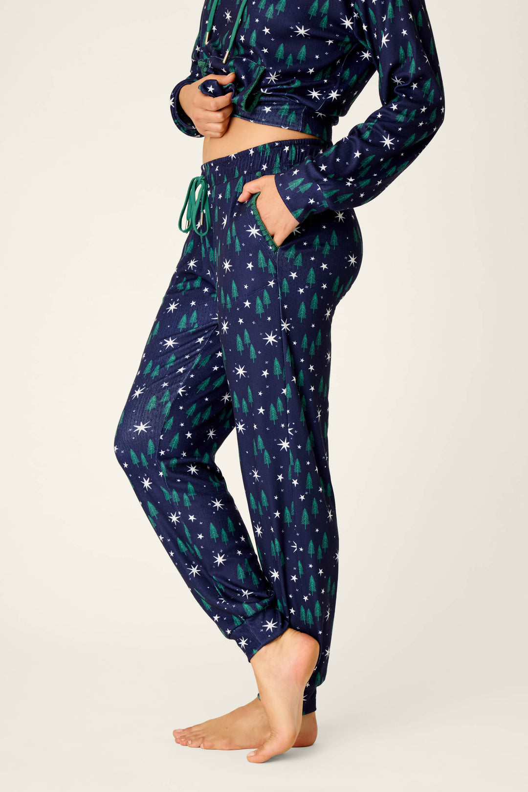 A person wearing the P.J. Salvage Under the Stars Magical Forest Print Joggers, featuring a navy blue background with white stars and green Christmas tree patterns, stands barefoot against a plain backdrop.