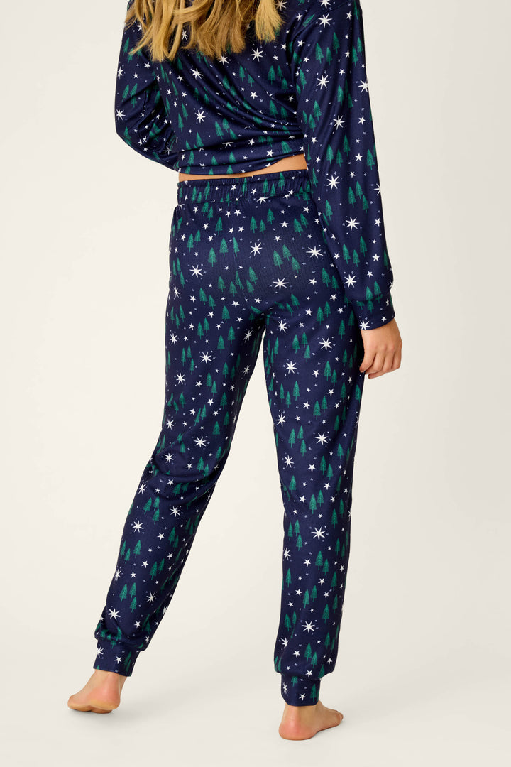 A person stands barefoot, shown from the back against a plain light background, wearing P.J. Salvage “Under the Stars Magical Forest Print Joggers” featuring a navy blue base with whimsical white star and green tree patterns.
