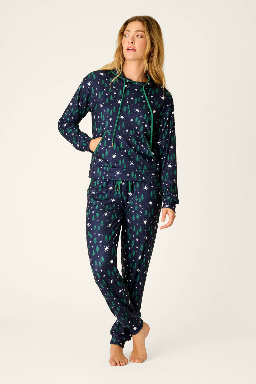A woman stands barefoot wearing the P.J. Salvage “Under the Stars Magical Forest Print” hoodie and jogger set, which features a matching navy blue design with green Christmas trees and white stars. With one hand in her pocket, she gazes to the side with a relaxed expression.