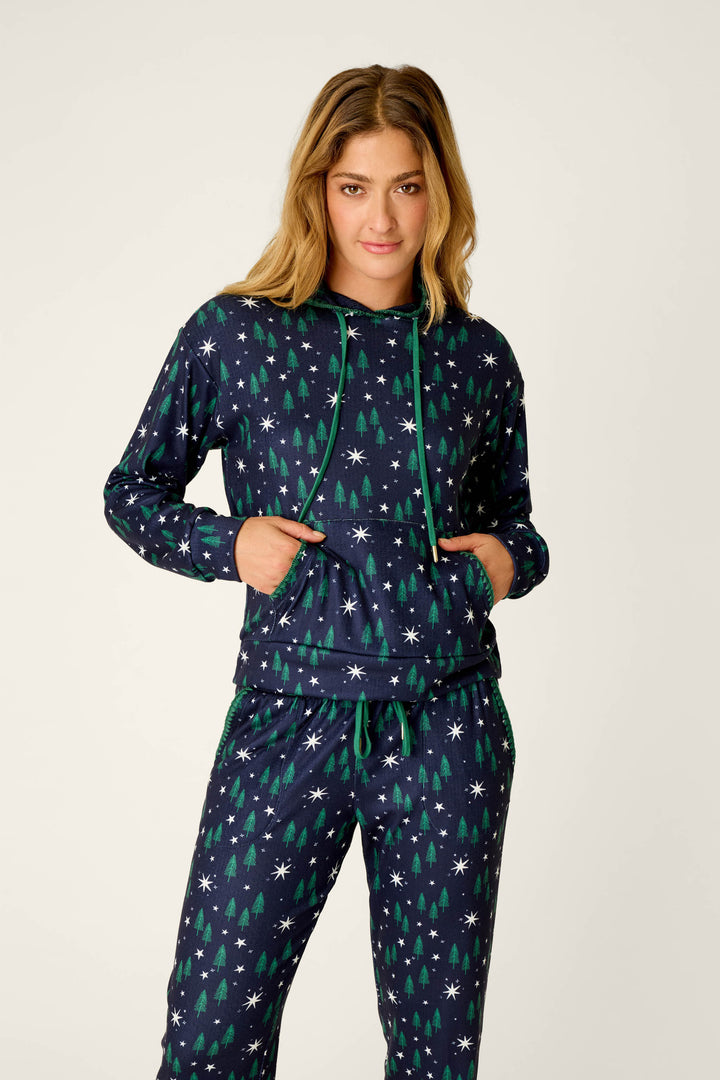 A person wearing the P.J. Salvage Under the Stars Magical Forest Print Hoodie & Jogger Set, featuring a navy background with green trees and white snowflakes, stands with hands in the hoodie pocket and looks at the camera.