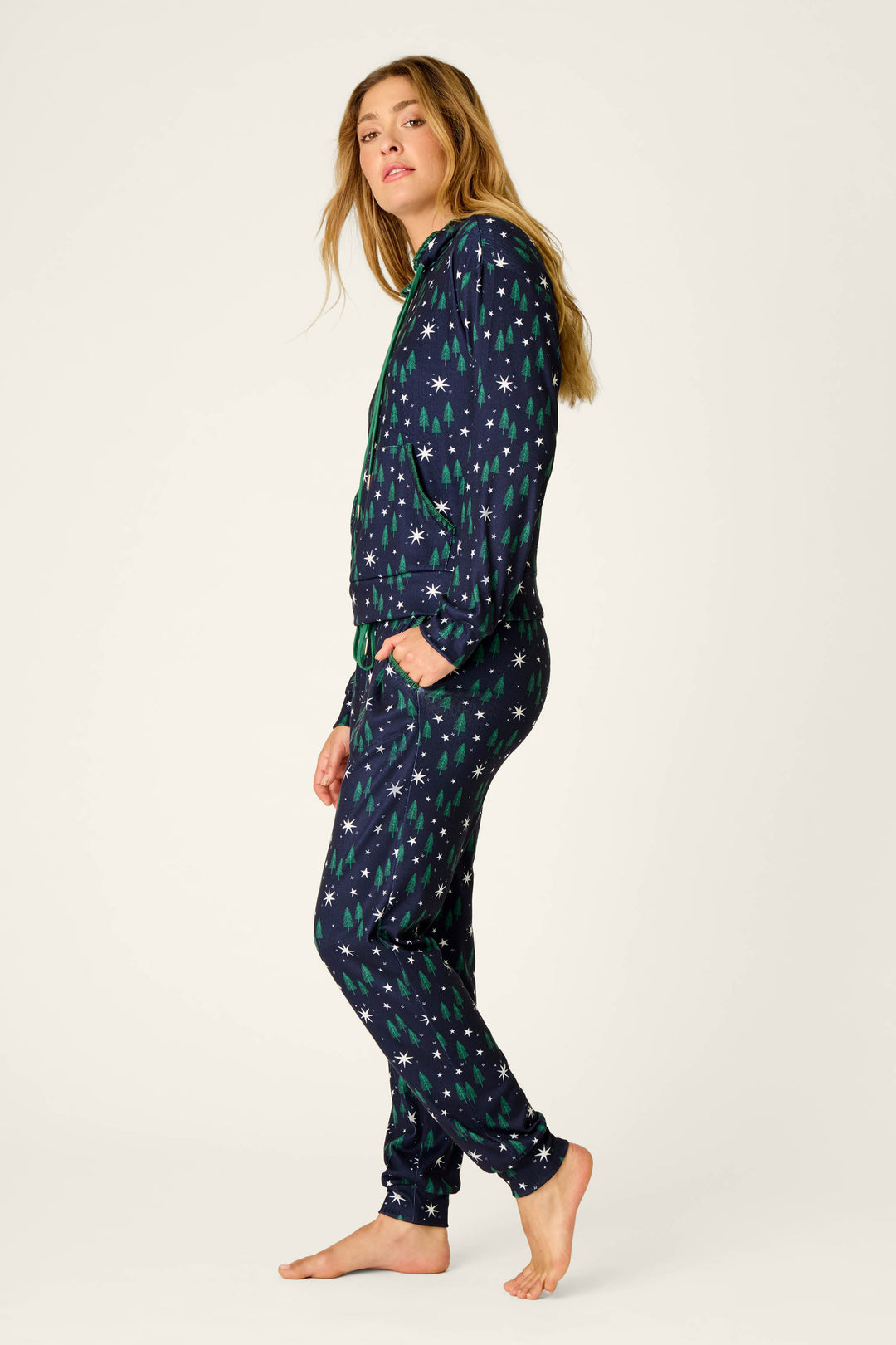 A person stands barefoot, wearing the P.J. Salvage “Under the Stars” Magical Forest Print Hoodie & Jogger Set—a dark blue, full-length loungewear ensemble featuring a green star pattern—while looking over their shoulder against a plain light background.