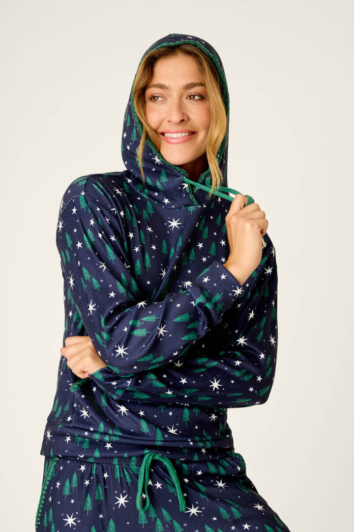 A woman smiles and looks to the side, wearing the P.J. Salvage “Under the Stars Magical Forest Print Hoodie & Jogger Set,” which features a navy design with green tree and white star patterns, as she holds the hoodie’s drawstring against a plain light background.