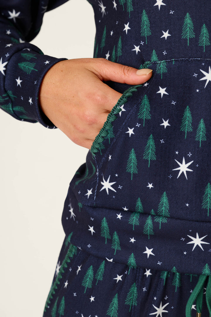 A hand is placed in the pocket of the Under the Stars Magical Forest Print Hoodie & Jogger Set by P.J. Salvage, featuring a navy blue design with green pine trees and white stars.