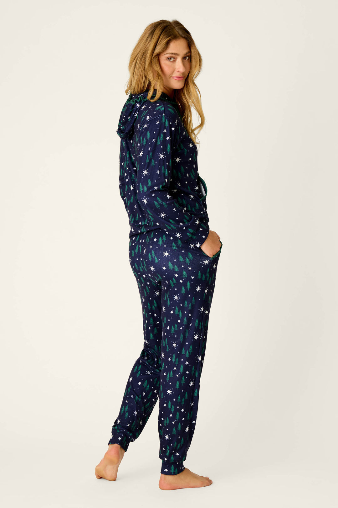 A woman stands barefoot against a plain background, hands in her pockets and looking over her shoulder, wearing the P.J. Salvage Under the Stars Magical Forest Print Hoodie & Jogger Set—a navy blue set adorned with a festive tree and star pattern.