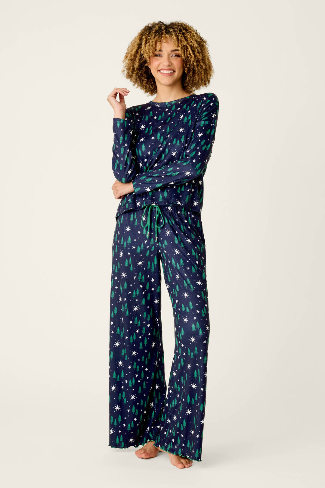 A person wearing the P.J. Salvage Under the Stars Magical Forest Print Long Sleeve Lounge Set, featuring a navy blue pajama set with green and white holiday tree and snowflake patterns, stands barefoot and smiles against a plain background.