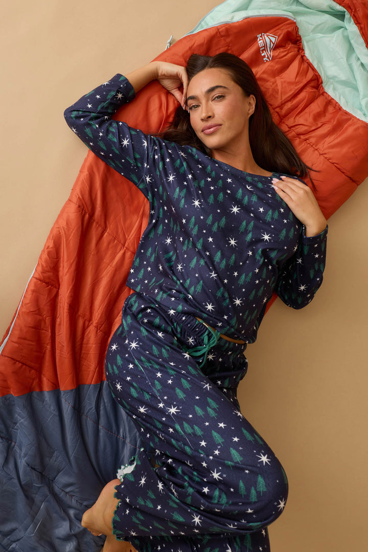 A woman wears the P.J. Salvage Under the Stars Magical Forest Print Long Sleeve Lounge Set as she lies on an orange and teal sleeping bag against a beige background, looking at the camera with one arm raised above her head.