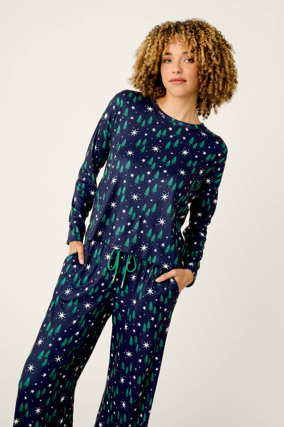 Wearing the “Under the Stars Magical Forest Print Long Sleeve Lounge Set” by P.J. Salvage, a person stands against a plain light background with hands in pockets, looking confidently at the camera. The navy blue set features whimsical green trees and white snowflakes in its print.