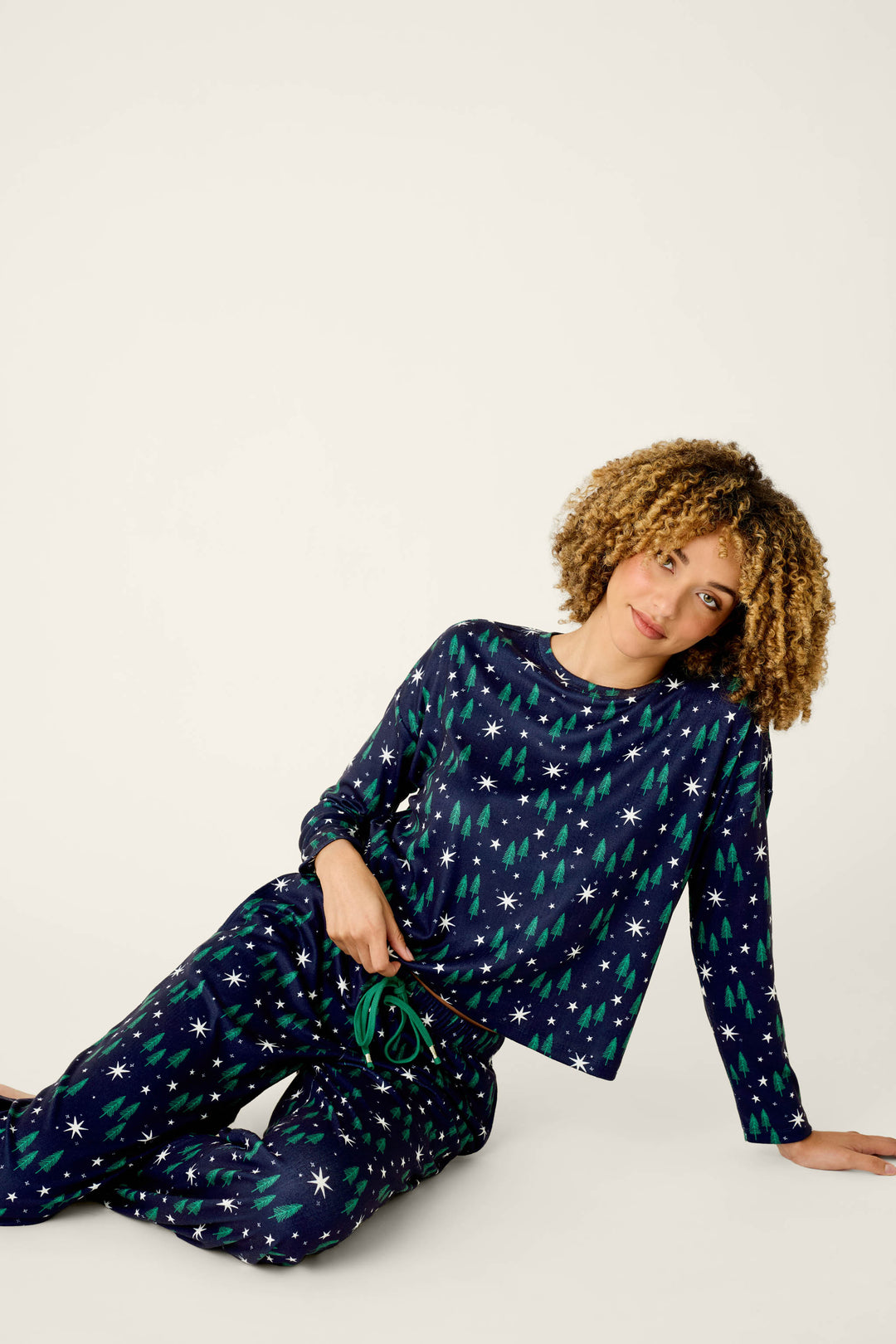 A person with curly hair sits on the floor against a plain background, wearing the P.J. Salvage Under the Stars Magical Forest Print Long Sleeve Lounge Set—navy pajamas adorned with green trees and white star patterns—while holding the drawstring of their pants and smiling slightly.