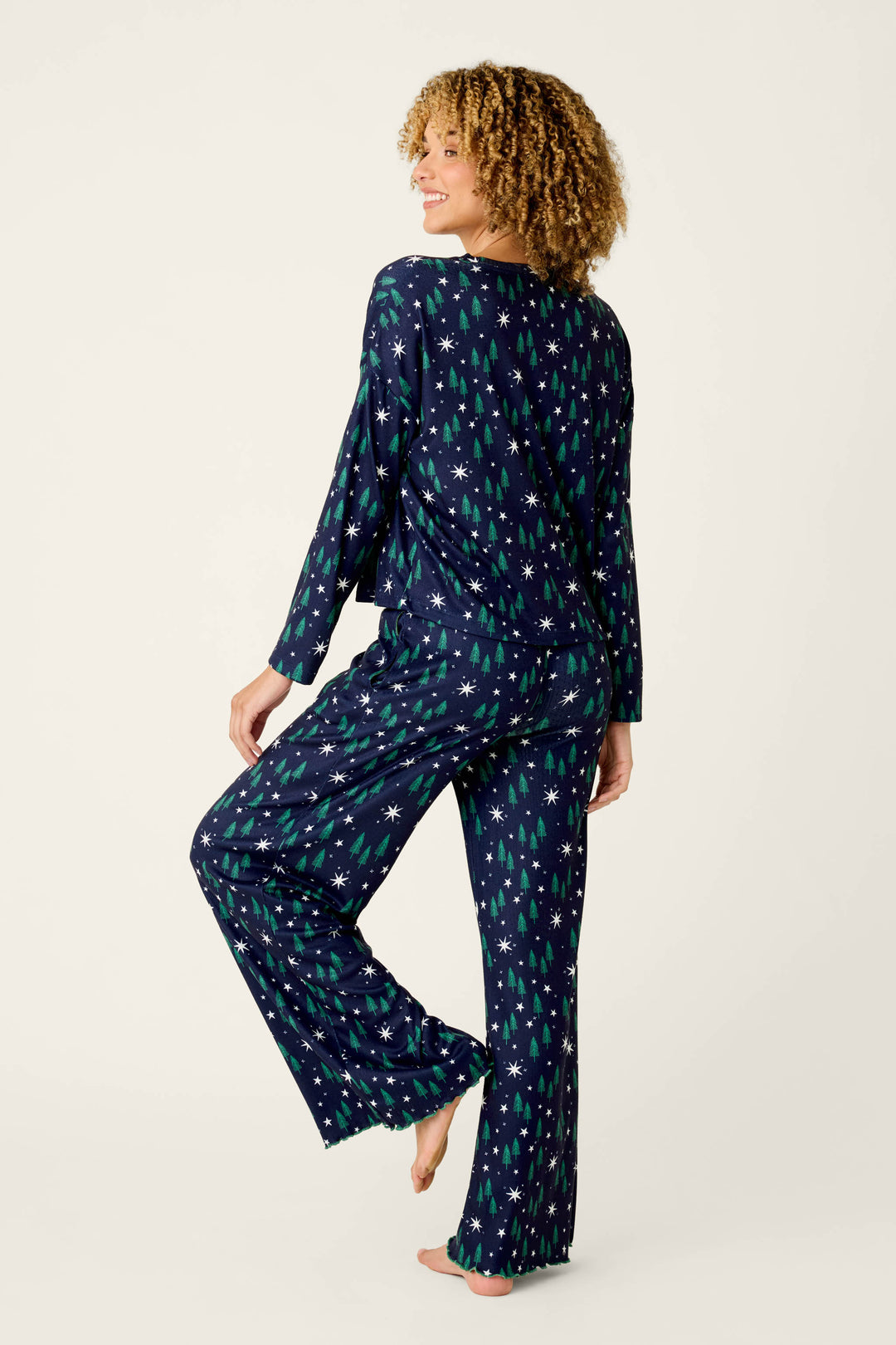 A person with curly hair models the P.J. Salvage “Under the Stars Magical Forest Print” long sleeve lounge set—navy pajamas adorned with green Christmas trees and white stars—standing barefoot and smiling over their shoulder against a plain light background.