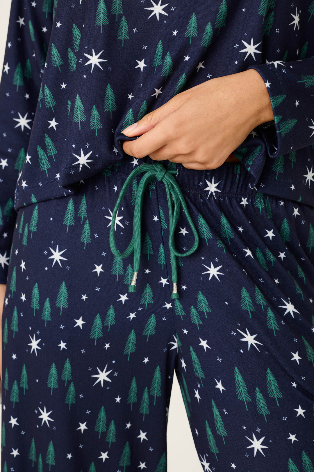 A close-up of a person wearing the P.J. Salvage Under the Stars Magical Forest Print Long Sleeve Lounge Set, featuring navy blue fabric adorned with green pine trees and white stars; one hand rests on the waistband, which is tied with a green drawstring.