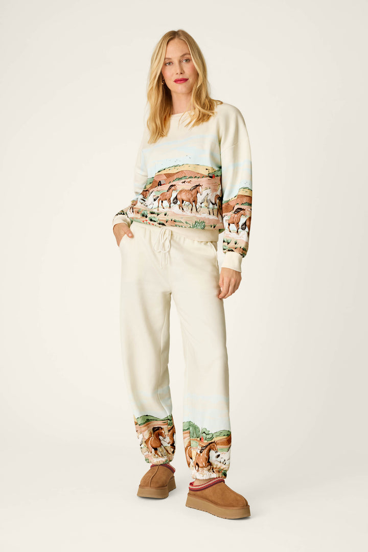 A woman with blonde hair and red lipstick stands facing forward against a plain white background, wearing the P.J. Salvage Western Ways Untamed Spirit Frankie Sweatshirt in cream—featuring horse and landscape prints—paired with matching sweatpants and brown slippers.
