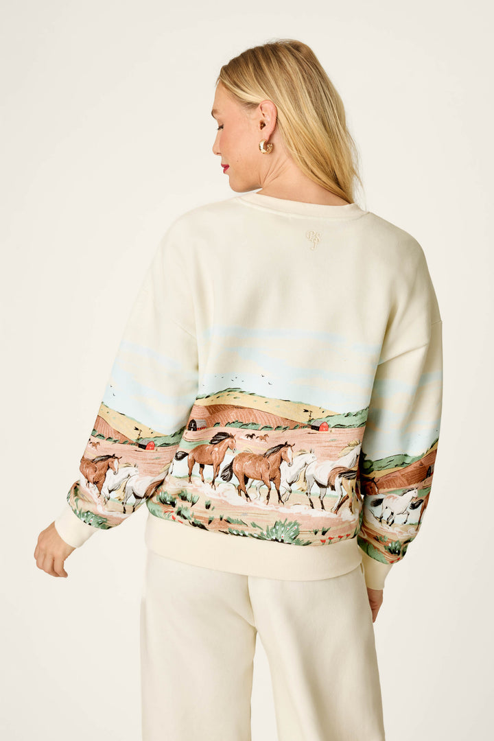 A woman with blonde hair models the P.J. Salvage Western Ways Untamed Spirit Frankie Sweatshirt, which showcases an illustrated desert scene with wild horses running across the back. She is photographed from behind to display the sweatshirt’s detailed design.