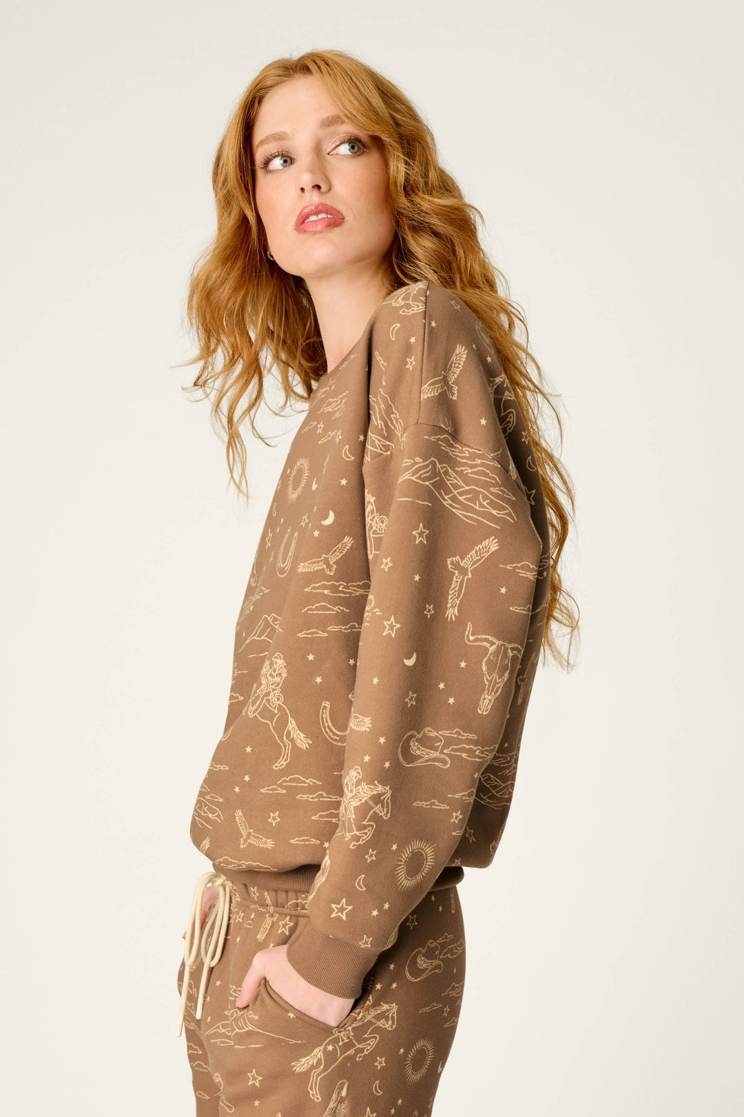 A woman with long, wavy red hair wears the P.J. Salvage Western Ways Ride ’Em Cowgirl Frankie Sweatshirt featuring beige celestial and animal prints as a matching set. She looks over her shoulder to the left against a plain light background.