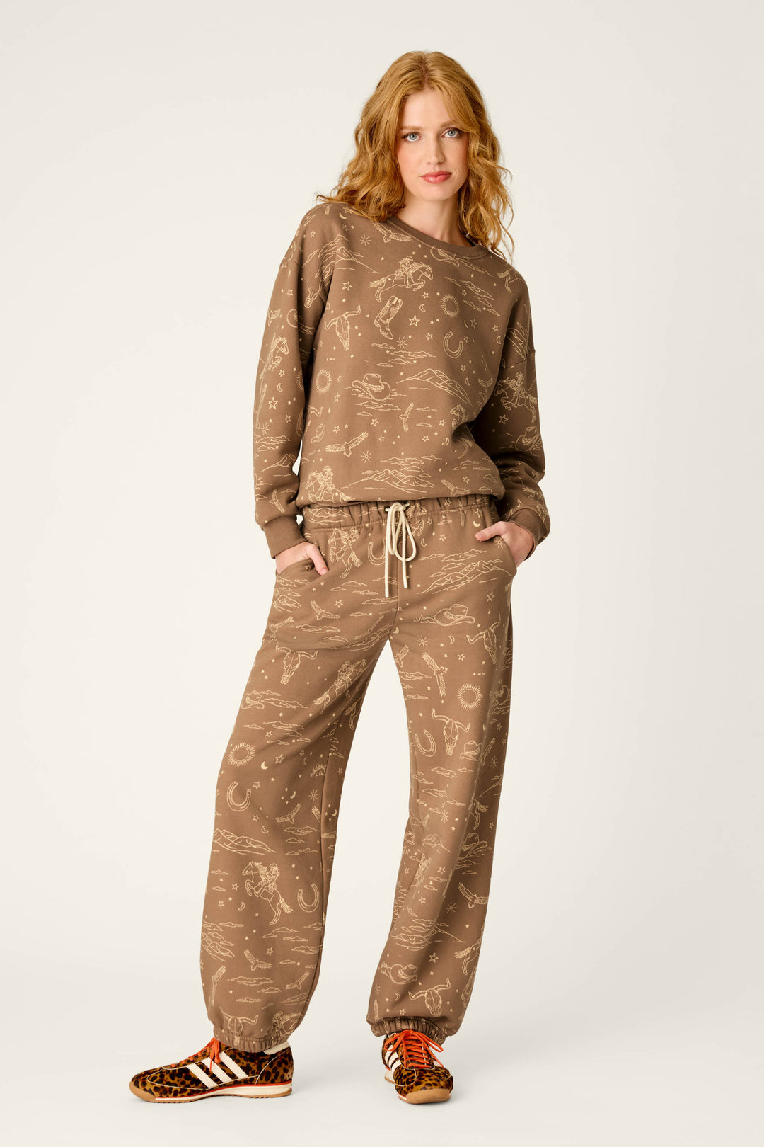 A woman with wavy red hair stands with her hands in her pockets against a plain background, wearing the P.J. Salvage Western Ways Ride ’Em Cowgirl Frankie Sweatshirt and matching sweatpants with a subtle print, paired with animal print sneakers featuring white stripes.