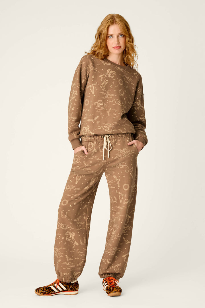 A woman with wavy red hair stands with her hands in her pockets against a plain background, wearing the P.J. Salvage Western Ways Ride ’Em Cowgirl Frankie Sweatshirt and matching sweatpants with a subtle print, paired with animal print sneakers featuring white stripes.