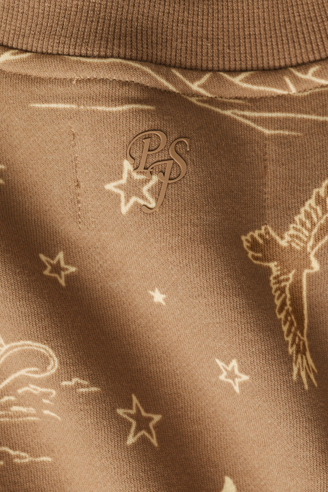 Close-up of the brown fabric of the P.J. Salvage Western Ways Ride ’Em Cowgirl Frankie Sweatshirt, featuring a subtle pattern of birds, stars, and lines, with an overlapping PS monogram at the center.