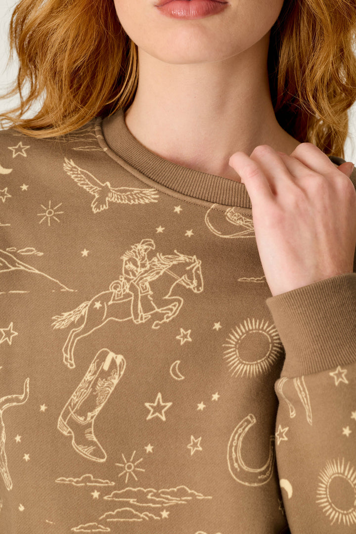 A woman wearing the Western Ways Ride ’Em Cowgirl Frankie Sweatshirt by P.J. Salvage, a brown sweatshirt featuring tan Western-themed line drawings like a cowboy on a horse, boot, stars, moon, horseshoe, eagle, and suns.
