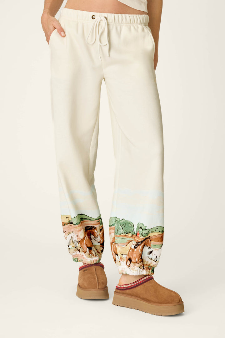 Wearing the P.J. Salvage Western Ways Untamed Spirit Good Hang Joggers, which feature a cream color, drawstring waist, and a colorful horse and landscape print near the ankles, paired with tan platform slippers.