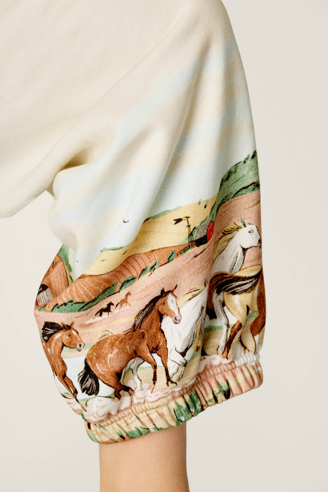A close-up of the Western Ways Untamed Spirit Good Hang Joggers by P.J. Salvage, featuring a cream-colored sleeve adorned with a vibrant print of running horses against a hilly landscape and red barn backdrop.