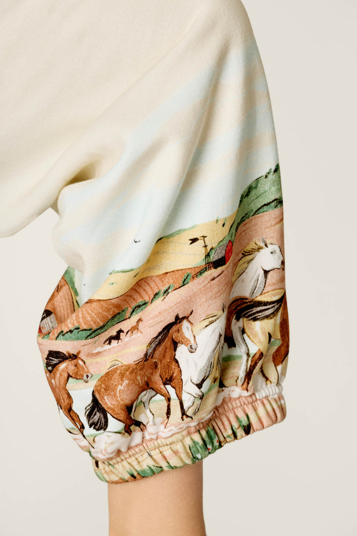 A close-up of the Western Ways Untamed Spirit Good Hang Joggers by P.J. Salvage, featuring a cream-colored sleeve adorned with a vibrant print of running horses against a hilly landscape and red barn backdrop.