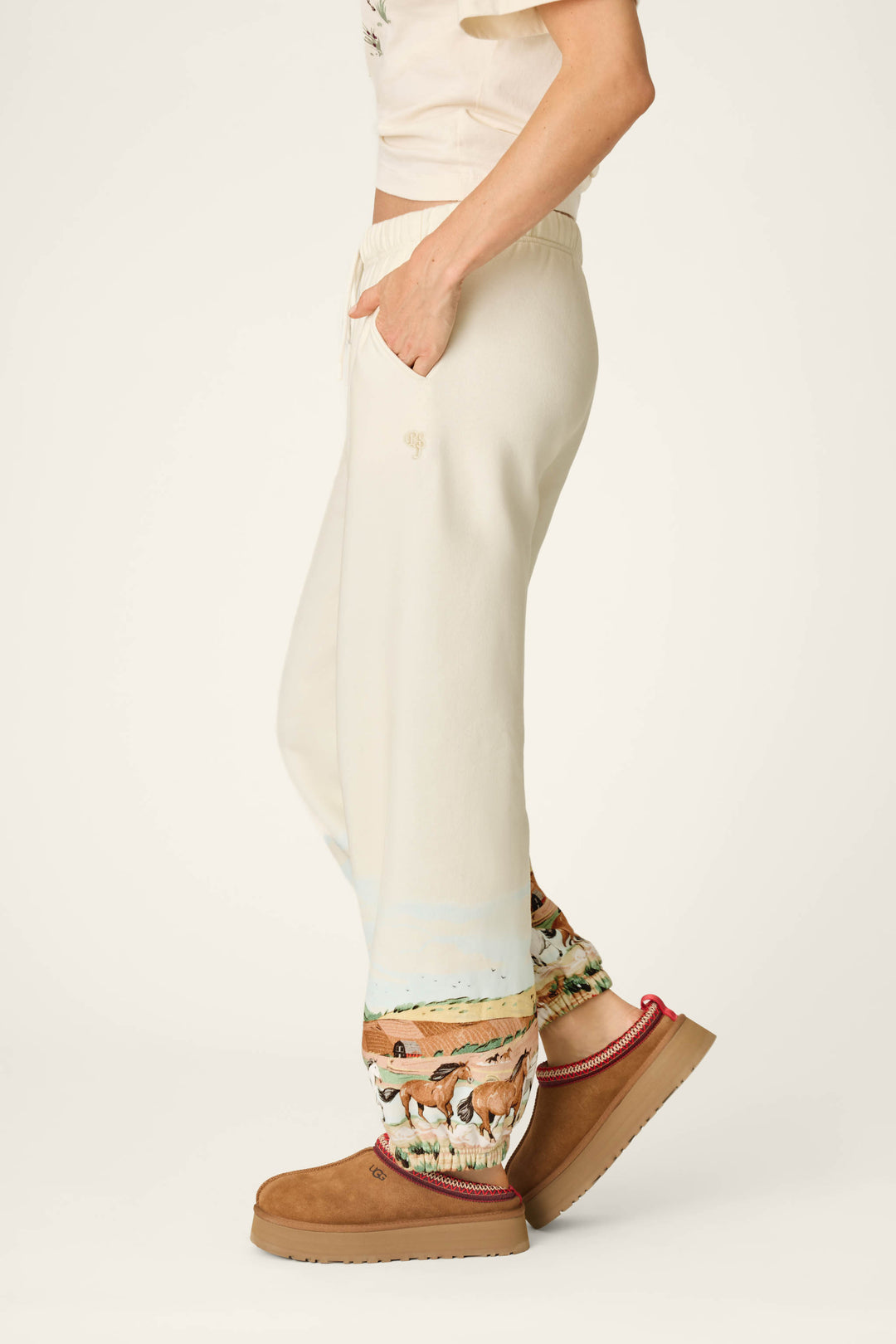 A person stands sideways with one hand in their pocket, wearing the P.J. Salvage Western Ways Untamed Spirit Good Hang Joggers, which feature a horse print near the ankles. They pair the joggers with tan platform slip-on shoes and a light top against a plain, light-colored background.