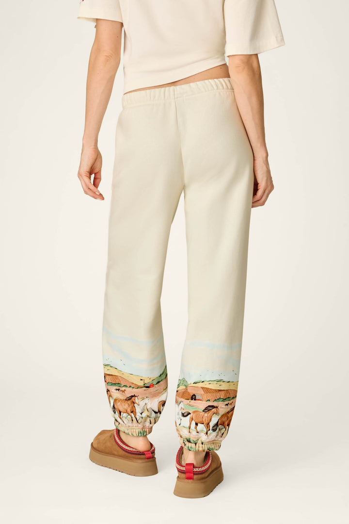 A person wearing the P.J. Salvage Western Ways Untamed Spirit Good Hang Joggers—cream pants featuring a landscape and horse design near the ankles—with brown shoes with red straps and a cream top, standing against a plain background.