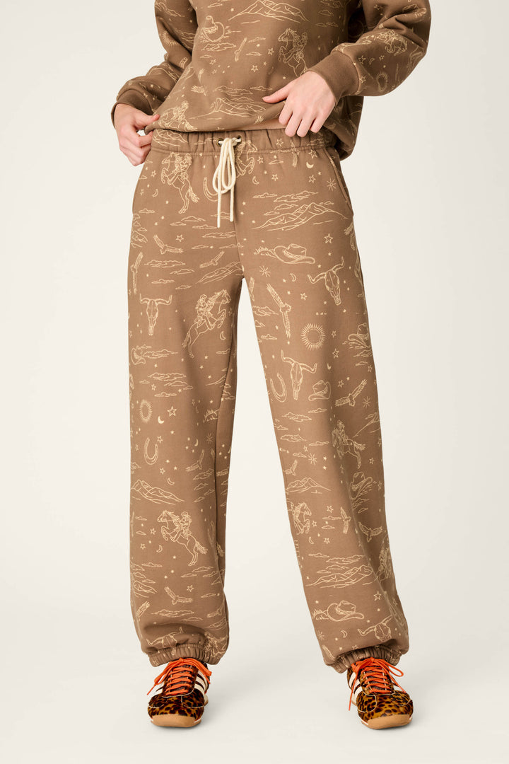 A person wears the P.J. Salvage Western Ways Ride ‘Em Cowgirl Good Hang Joggers, featuring a light tan Western-themed pattern, paired with a matching sweatshirt and orange leopard-print sneakers. Their hands are tucked into the joggers’ waistband, standing against a plain, light-colored background.