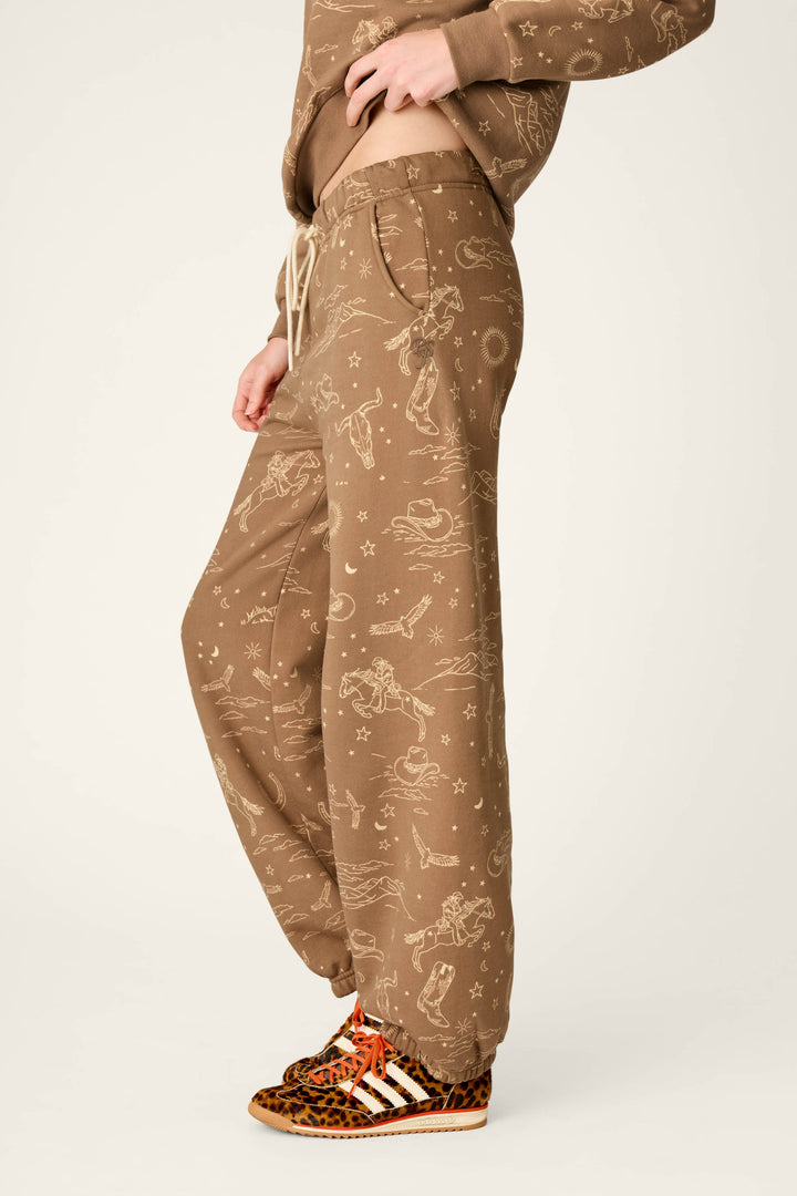 A person wearing the P.J. Salvage Western Ways Ride ’Em Cowgirl Good Hang Joggers, featuring light-colored constellation and cosmic patterns, with a matching top and brown leopard print sneakers with white stripes, standing sideways against a plain background.